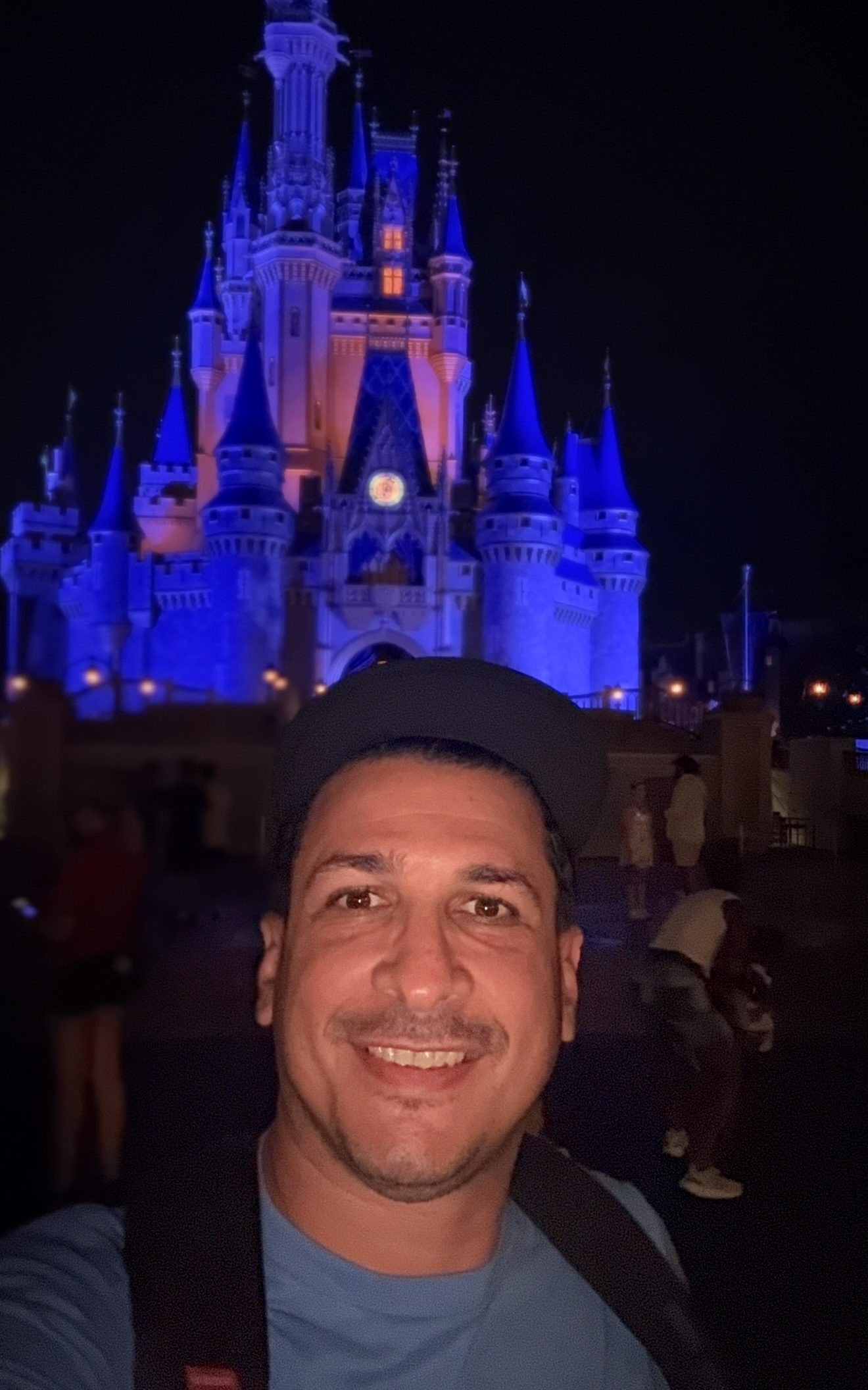 Hombre sonriendo con una camiseta azul en primer plano, frente a un castillo iluminado de noche, con luces azules y moradas.